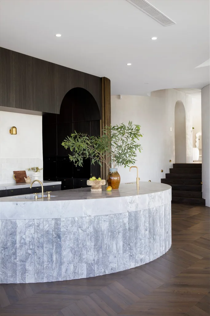 Luxurious kitchen with curved marble island, brass fixtures, a vase filled with greenery, set on dark herringbone flooring. |