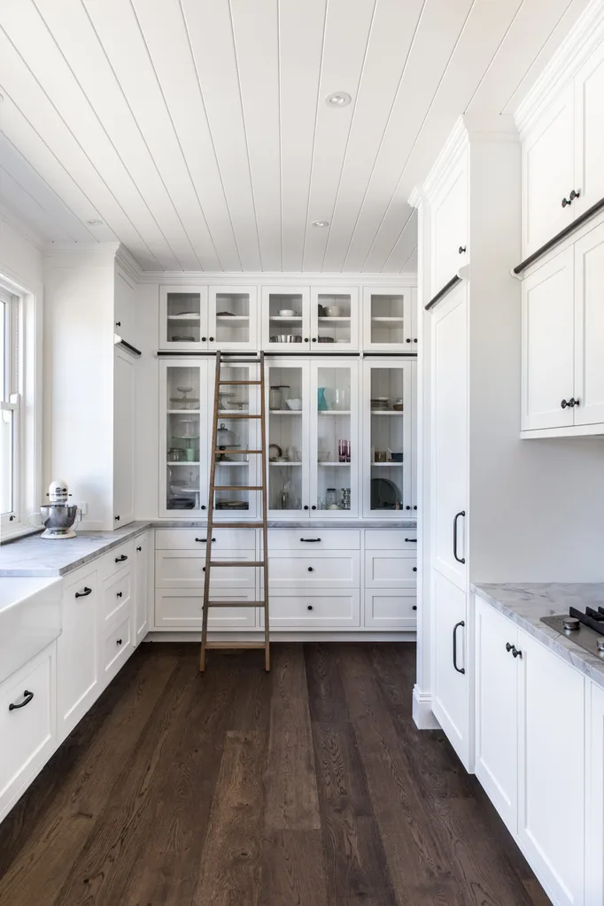 Bright and spacious butler's pantry with white cabinetry, glass-front cabinets, dark wood floors, and a wooden ladder for reaching upper shelves. The hardware is a dark metal, which complements the dark tone of the floor.