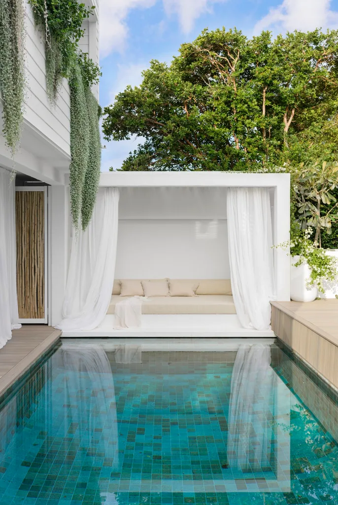 Outdoor pool area with white cabana, beige cushioned seating, hanging foliage, and a lush green tree background.