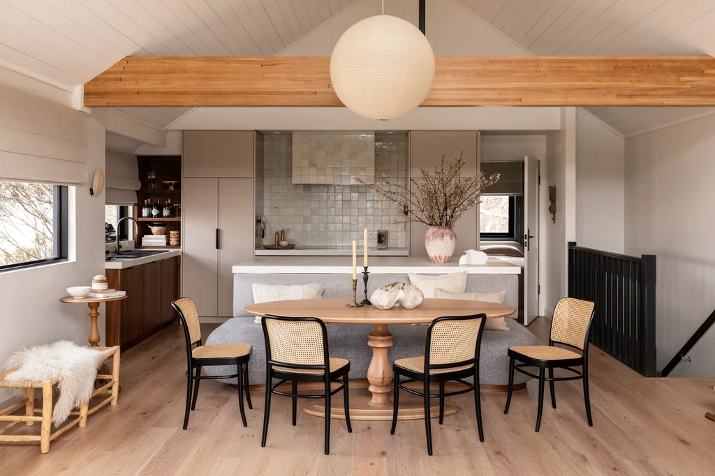 Open-plan kitchen and dining area with an oval wooden table, wooden chairs, neutral tones, a large pendant light, and modern decor. The ceiling is vaulted with one wooden beam exposed.