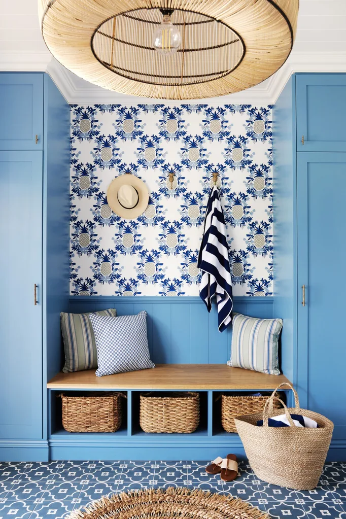 Coastal-themed mudroom with blue cabinets, floral wallpaper, hats, striped towels, cushions, wicker baskets, and a rattan light. A built-in bench seat is made from a light timber tone, with wicker baskets underneath and this breaks up the blue tones of the space.