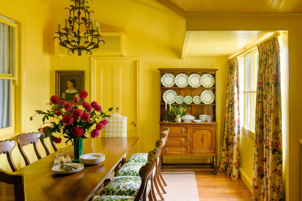 Colour drenched yellow dining room with a wooden table, wooden dining chairs with seat cushions that have a floral pattern, chandelier, flowers, and a wooden cabinet with decorative plates.