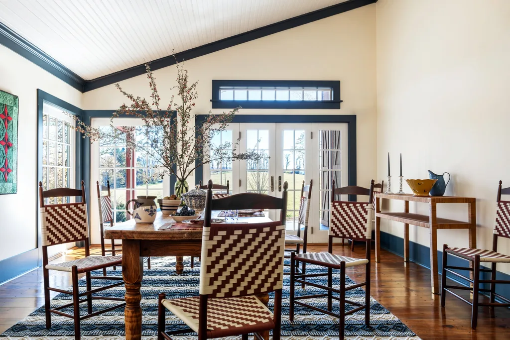 Rustic dining room with woven chairs, wooden table, decorative pottery on the dining table and a wooden side table, and large windows overlooking a garden. The skirting board, cornices and and trim around the windows and doors is painted in dark blue. The rug underneath the table is blue and white.
