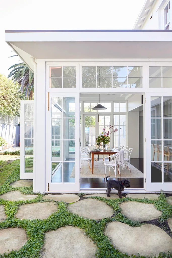 Open French doors leading to a bright dining area; table set with a vase, white chairs. In the foreground a small dog stands on a stone path around the exterior side of the house.