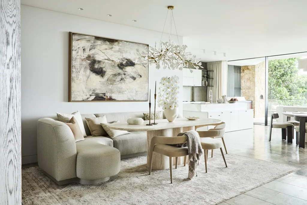 Luxurious dining room with a plush sofa, abstract art, modern chandelier, and dining table in a bright, airy space. The plush sofa is acting as banquette seating against the wall for the dining table, with three dining chairs on the opposite side.
