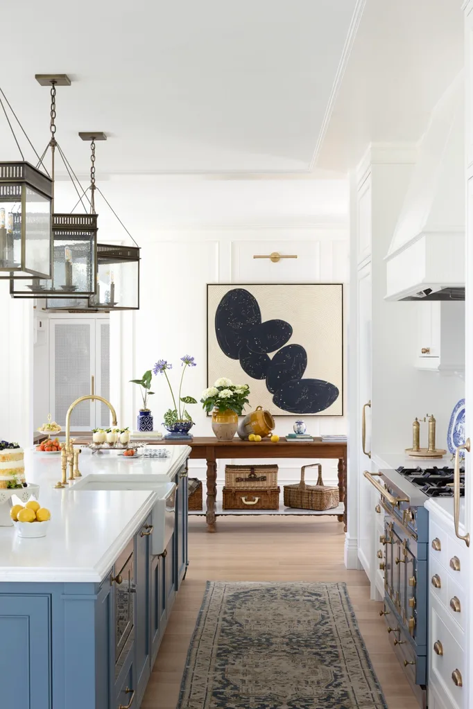 Elegant kitchen with blue shaker style joinery, white shaker style joinery, gold pulls and handles on drawers and cupboards. The kitchen island has three large pendants above it. Fruit is displayed on the island. In the background, a hallway has a large abstract painting on the wall and a wooden hallway table with flowers decorating it.