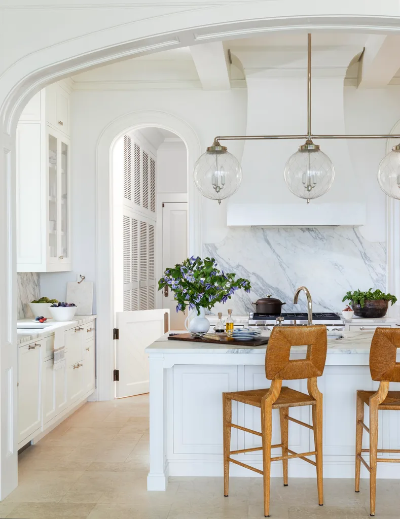 Bright kitchen with a marble island and splashback, wicker and wooden bar stools, glass and chrome pendants above the island, and a vase with lilac flowers in it. Cabinetry is shaker style and an arch leads to a butler's pantry.