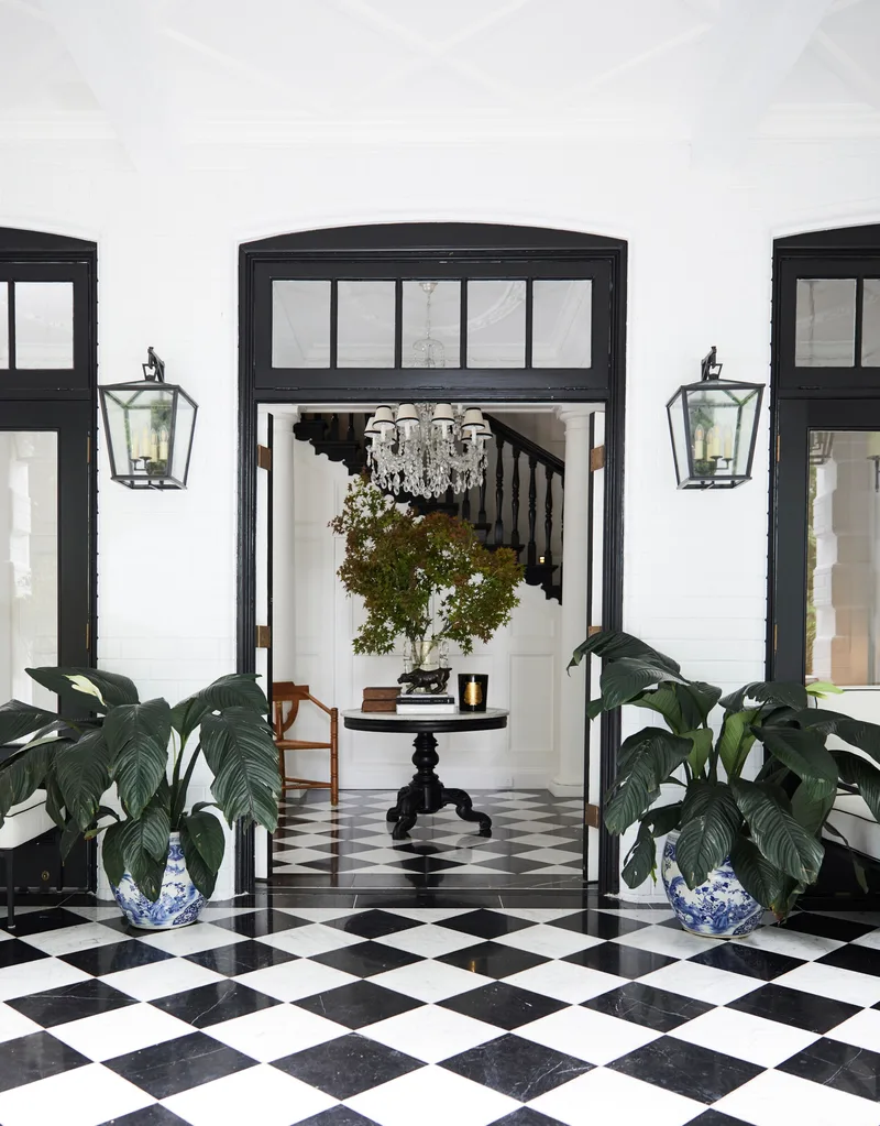 Grand foyer with black and white checkerboard tile floor, large plants, and chandelier. Two lanterns are positioned on the wall on either side of the open door.