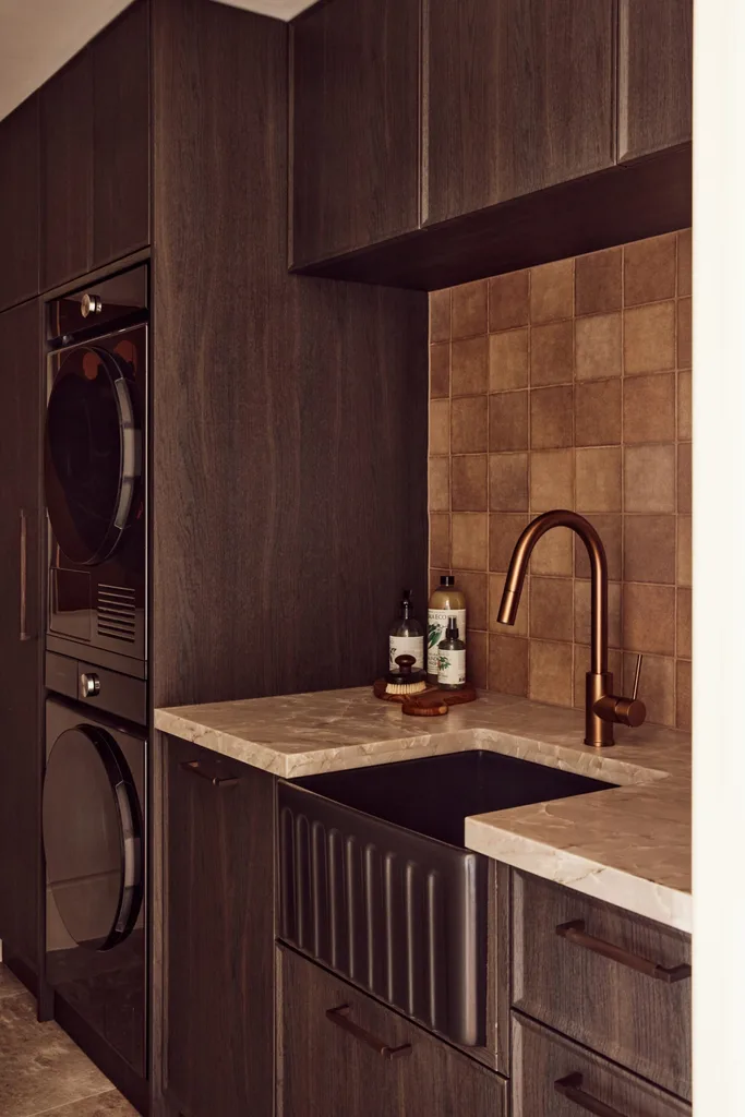 A laundry laundry with dark wood cabinetry, stacked washer and dryer set in to the cabinetry, bronze faucet, and square tiled splashback. The tiles are shades of cream, browns and other neutral tones.