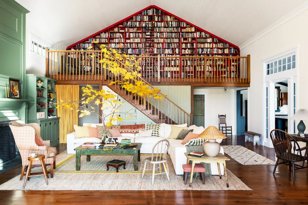 Spacious living room with a white sofa, vibrant cushions, autumnal decor, and a large bookshelf on the upper floor. The ceiling is pitched and the bookshelf on the upper floor is built into the wall, following the pitched roof line. A wooden ladder is set up against the bookshelf.