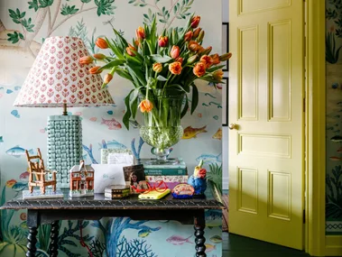 Decorative table with lamp, tulips, books, and ornaments. Yellow door and nature-themed wallpaper in the background.