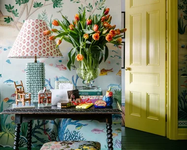 Decorative table with lamp, tulips, books, and ornaments. Yellow door and nature-themed wallpaper in the background.