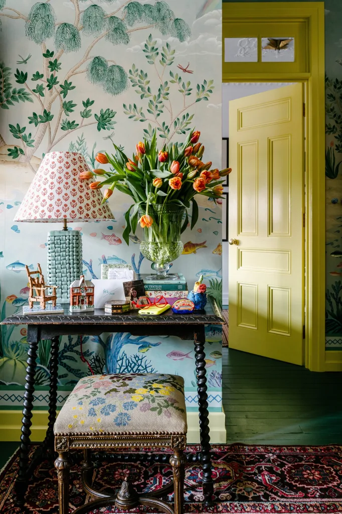 Decorative table with lamp, tulips, books, and ornaments. Yellow door and nature-themed wallpaper in the background.