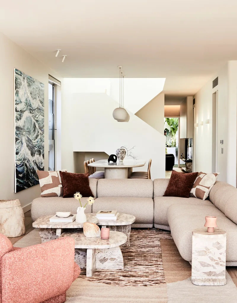 Darren Palmer's Bondi home living room with cream couch and pink armchair. The couch is L-shaped with a large painting of an ocean with waves. A dining area is in the background. The coffee table in the living room is made from marble.