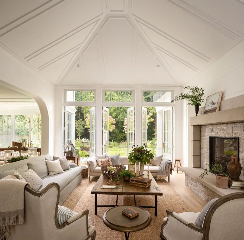 Elegant living room with vaulted ceiling, large windows opening to a garden, stone fireplace, and cozy seating arrangement. |The armchairs, sofa and cushions are all creamy neutrals in tone, arranged around a sisal rug and wooden coffee table.
