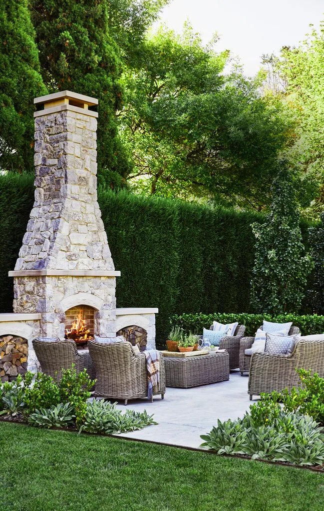 Outdoor living area with tall stone fireplace, wicker furniture, and lush greenery under a clear sky. |