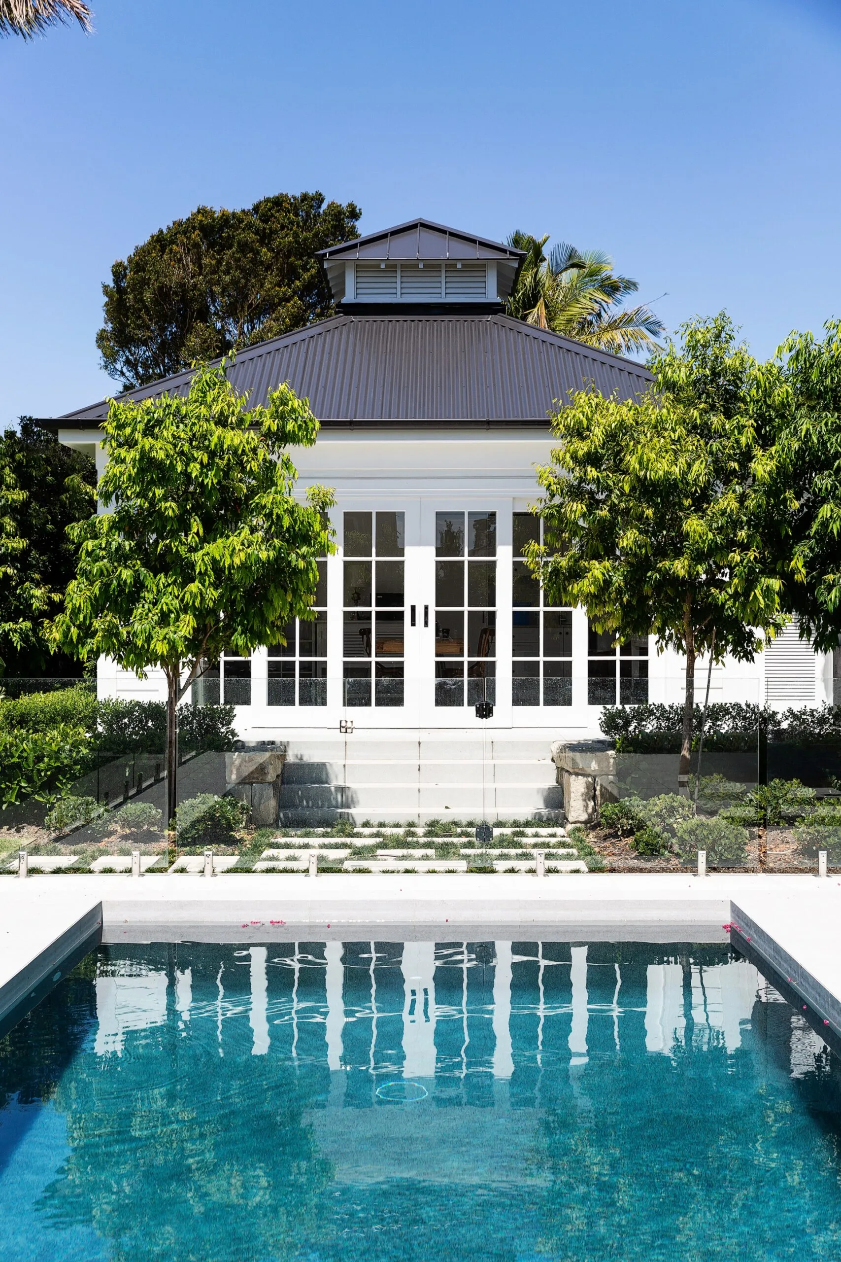 Large pool house with white French doors, surrounded by greenery, with a swimming pool in the foreground under clear blue sky.