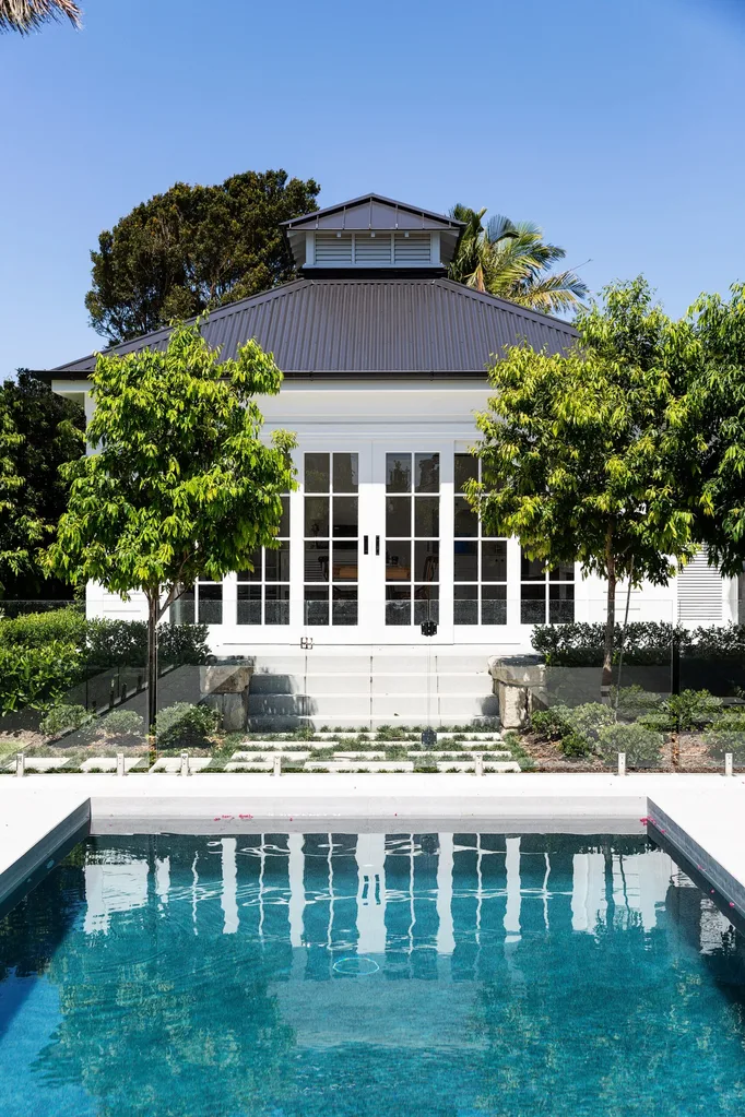 Large pool house with white French doors, surrounded by greenery, with a swimming pool in the foreground under clear blue sky.