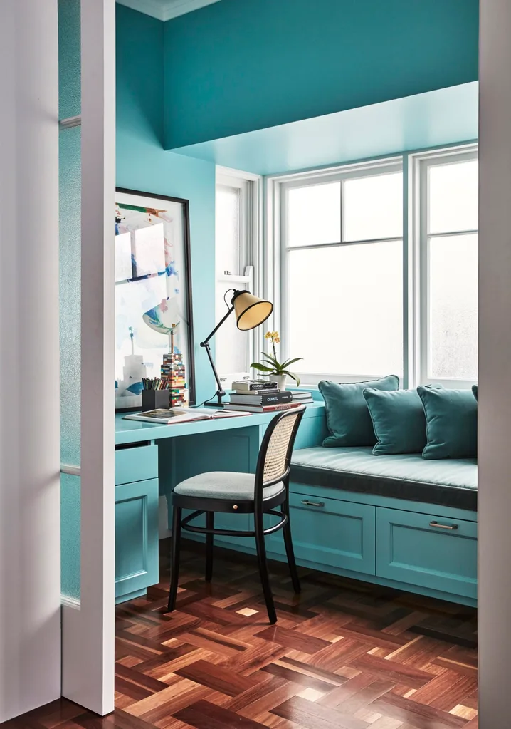Cozy home office with a teal color scheme, desk, chair, books, lamp, and window seat with cushions. The floors are timber and the teal shade colour drenches the room, covering the walls, built-in desk and window seat. Even the cushions on the window seat are in a teal shade.