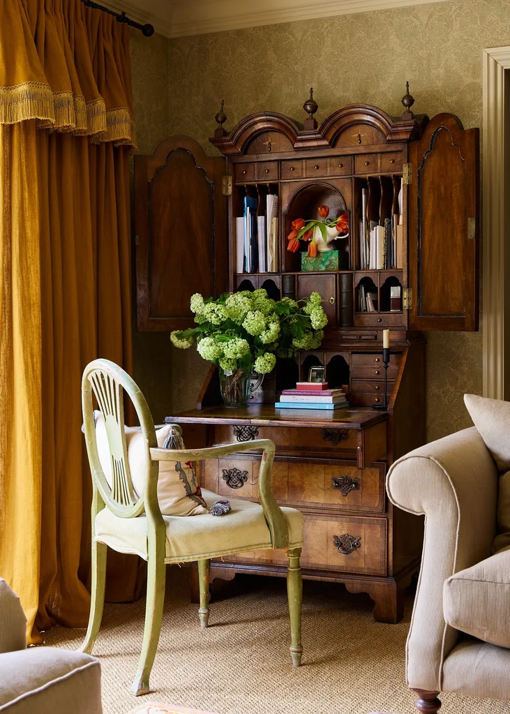 Antique wooden desk with open hutch, green dining chair with cushions, vibrant flowers and books on the desk, in cosy room with gold curtains, including a fringed valance. Neutral toned wallpaper is on the walls and neutral toned carpet is the flooring.
