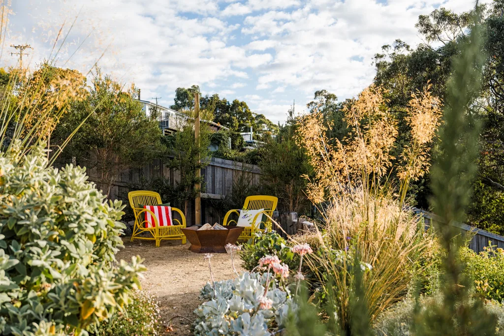 A backyard area with native flowers, two yellow outdoor chairs and a steel fire pit.