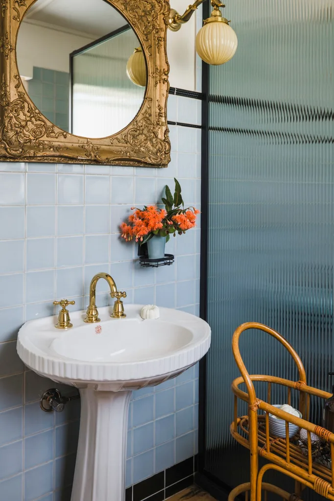 A bathroom with a pedestal basin with gold tapware, underneath a mirror with a gilded frame. The walls feature square blue tiles and a dado rail of dark blue tiles.
