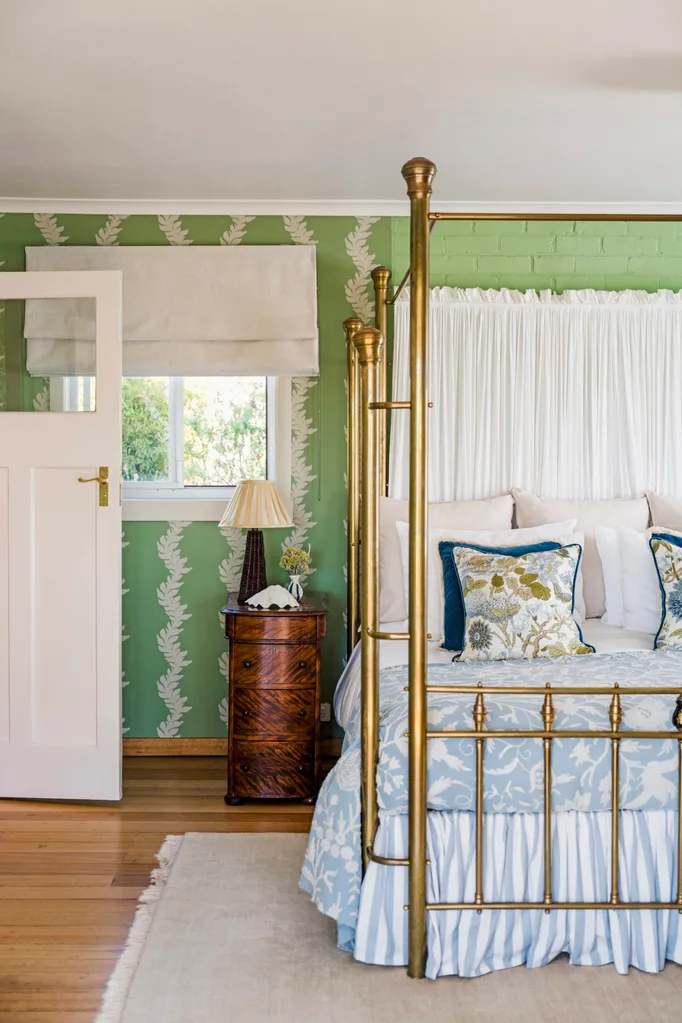 A bedroom with green wallpaper, neutral Roman blinds and a timber side table positioned beside a four-poster bed. The four-poster bed has a gold frame and the bedlinen is a mix of light blues, whites and dark blues.