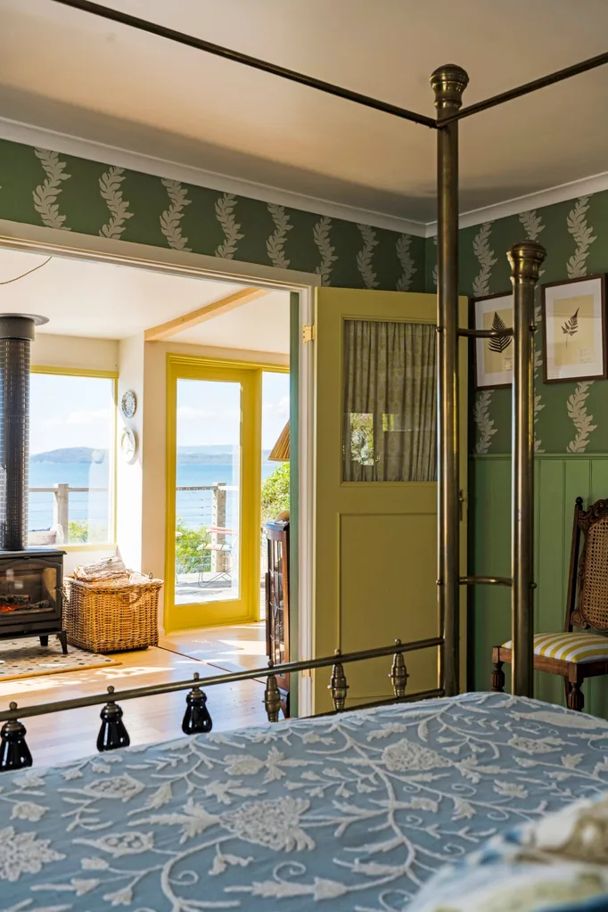 The four-poster bed is in the foreground of this photo, with a golden frame and blue and white bed linen. The walls of the bedroom are green VJ-panelled wainscoting and green wallpaper. Doors open to the fireplace in the living room, with sea views visible beyond.