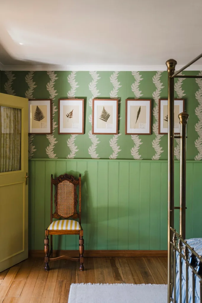 The corner of the four-poster bed is visible in this photo. The bedroom wall has green VJ-panelled wainscoting with green and cream patterned wallpaper above it. Five botanical prints are framed and placed atop the wallpaper.
