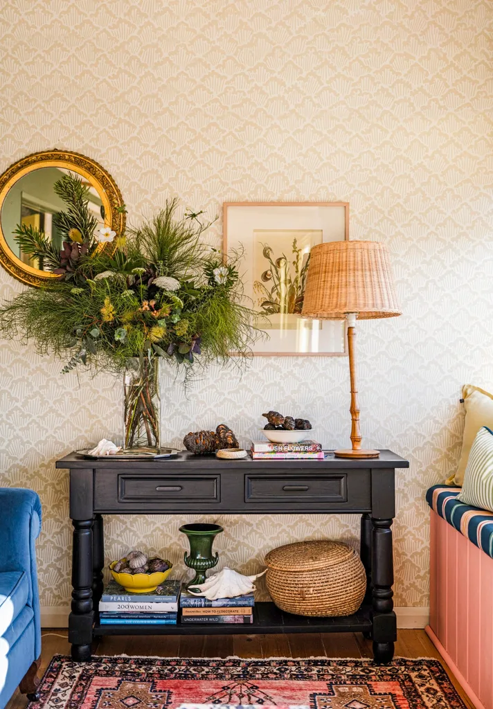 A console table is positioned in front of a wall covered in a neutral toned wallpaper. A vase with fresh flowers is on top of the console table, in between a round, gilded mirror and a botanical illustration. A rattan table lamp is on the other side of the console table.