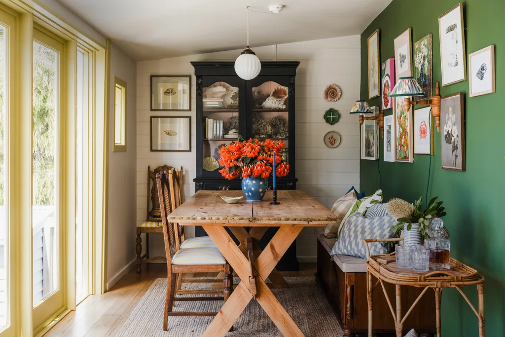 The dining room in this Tasmanian Airbnb features white VJ-pamelled walls, beside a green feature wall. This green feature wall has 10 artworks hung on it, with two wall sconces. The dining table is timber, with two dining chairs with timber frames and a banquette on the opposite side, decorated with cushions.
