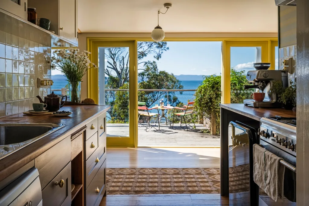 The kitchen of this Tasmanian airbnb has a U-shaped layout, a square-tiled splashback and a small fridge beside the cooker and stovetop. From the kitchen, looking to the back of the house you can see the deck and the sea views beyond.