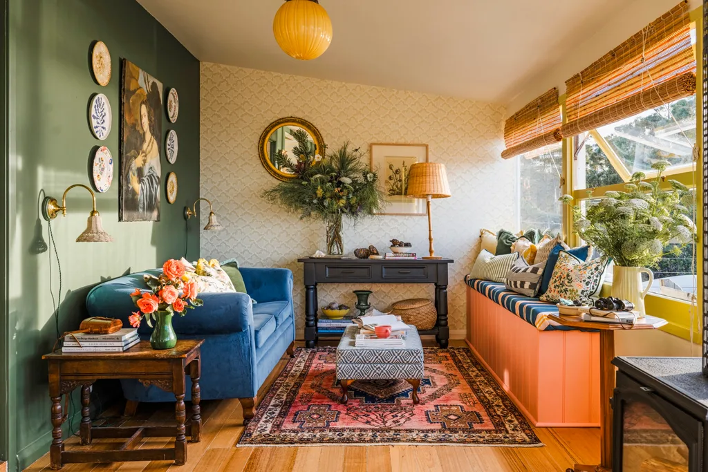 The living room of this Airbnb in Tasmania is filled with colour and pattern. A green wall is decorated with ceramic paints and a large oil painting of a woman. A blue couch is positioned opposite to a window seat, upholstered in a striped fabric and decorated with plentiful cushions. An ottoman doubles as a coffee table on top of a rug.