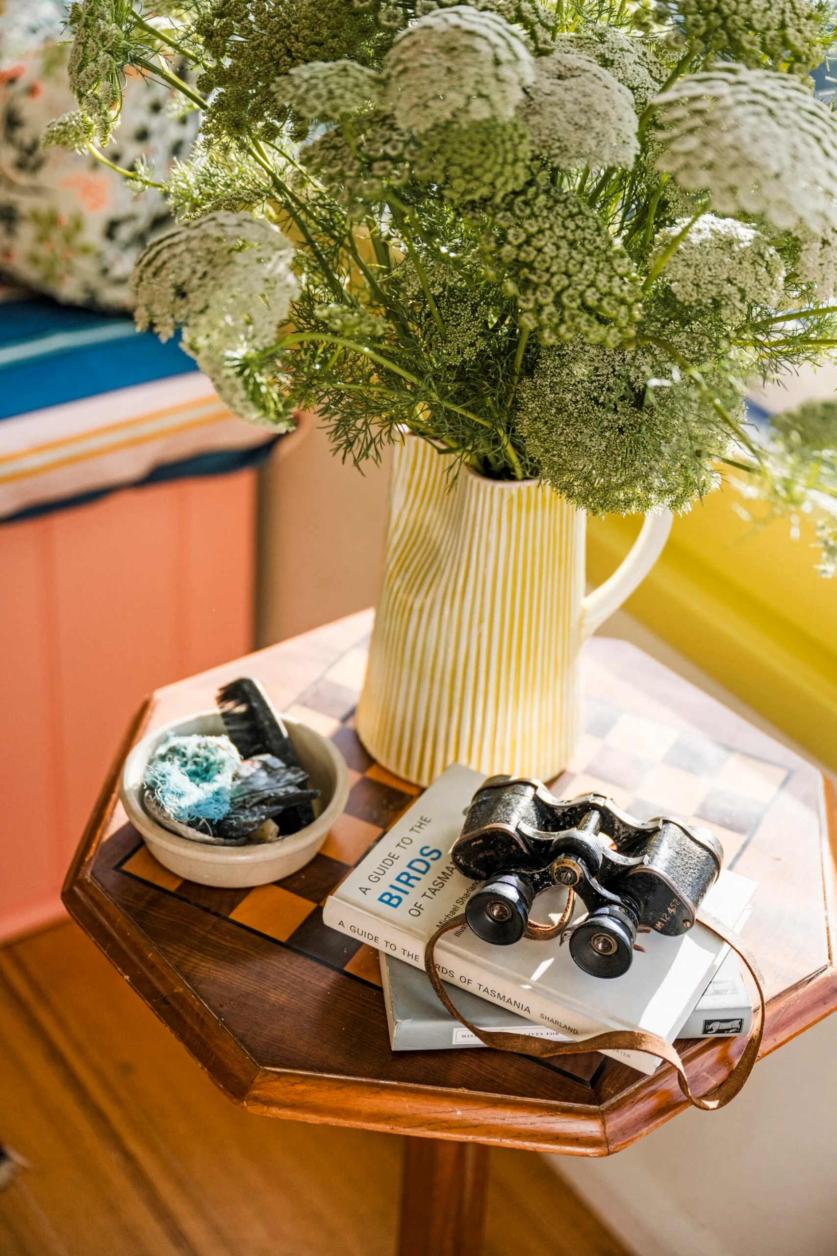 A close up of a side table that was an antique chessboard table. A yellow and white striped jug is filled with flowers, positioned beside antique binoculars and a book about Tasmanian birds.