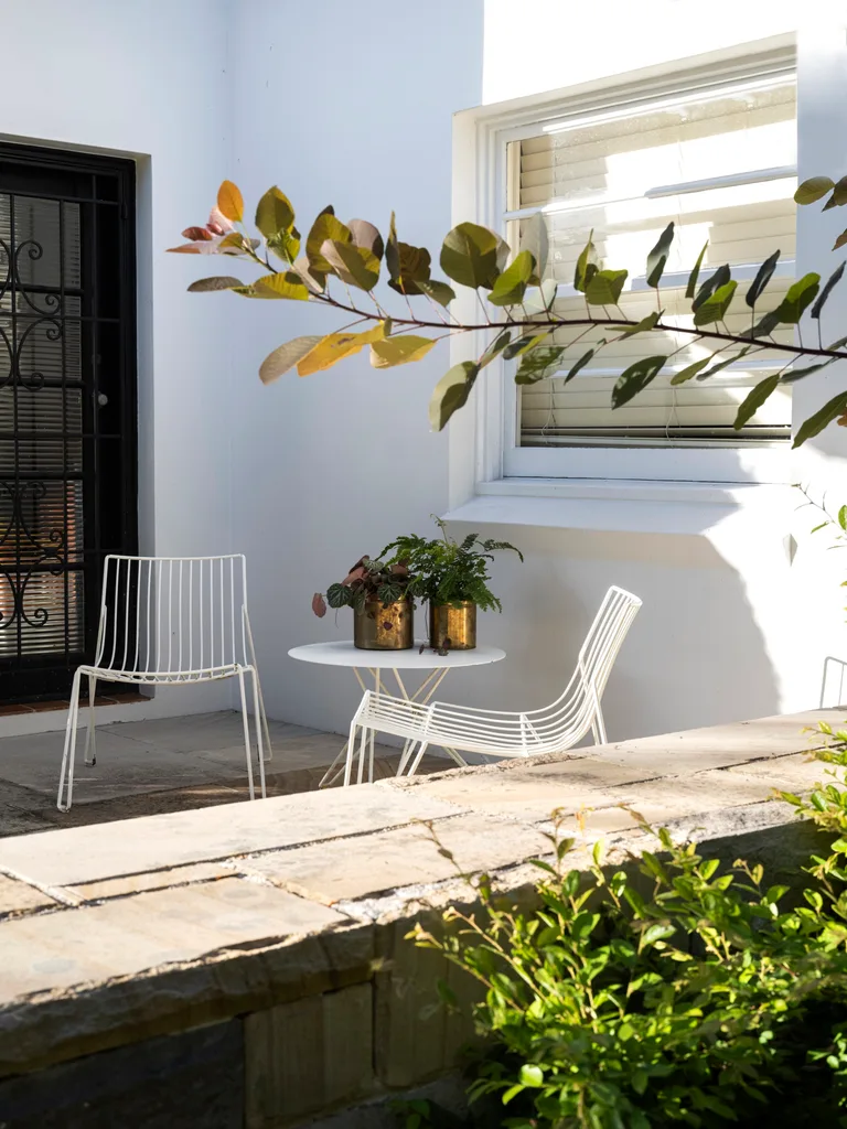 The front terrace of the home has white walls and white outdoor furniture. The furniture is a white metal and is comprised of two chairs and a round table.