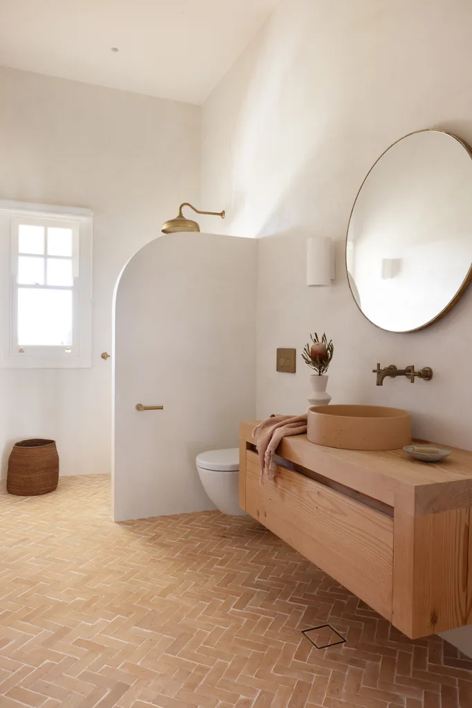 Minimalist bathroom with wooden vanity, round mirror, walk-in shower, and terra-cotta floor tiles.