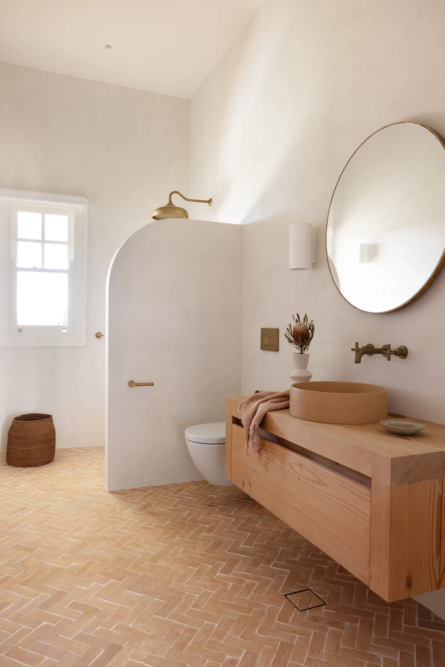 Minimalist bathroom with wooden vanity, round mirror, walk-in shower, and terra-cotta floor tiles.