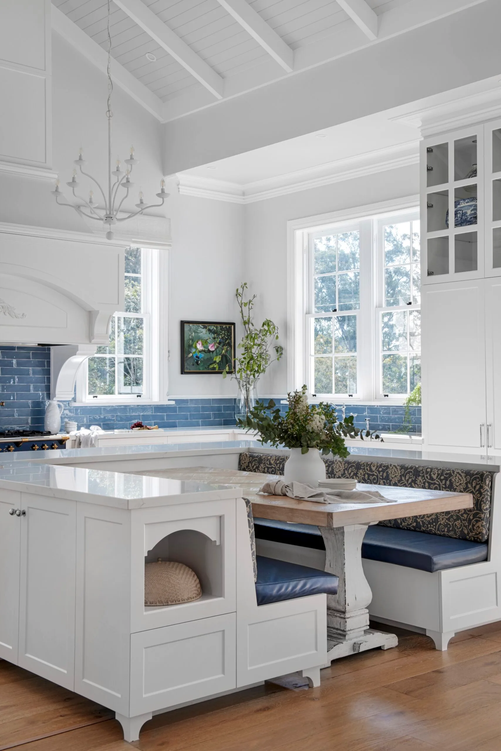 Bright kitchen with white cabinets, blue tile splashback, a white chandelier, and a cozy breakfast nook with blue cushions. The breakfast nook has a dining table positioned in the middle of two built-in banquette seats. A vaulted ceiling is finished in white with exposed beams and VJ panelling.
