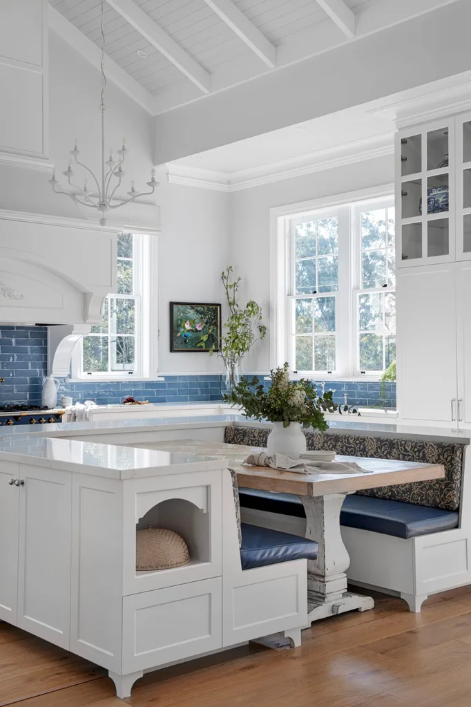 Bright kitchen with white cabinets, blue tile splashback, a white chandelier, and a cozy breakfast nook with blue cushions. The breakfast nook has a dining table positioned in the middle of two built-in banquette seats. A vaulted ceiling is finished in white with exposed beams and VJ panelling.