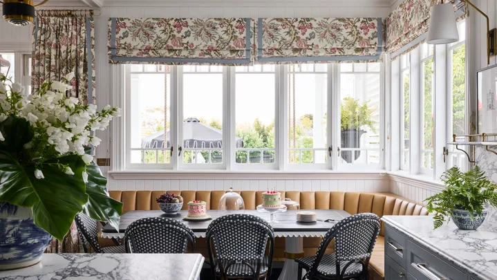 Elegant kitchen with a breakfast nook in front of the kitchen island. This is built around the corner of the room, underneath windows with floral Roman blinds. A dining table is surrounded on two sides by built-in banquette seating, with dining chairs on the other two sides. Marble countertops are visible in the foreground and the dining table is set with cakes and fruit.