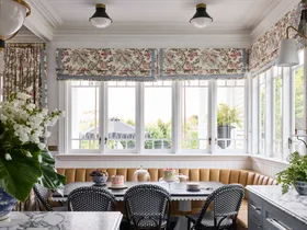 Elegant kitchen with a breakfast nook in front of the kitchen island. This is built around the corner of the room, underneath windows with floral Roman blinds. A dining table is surrounded on two sides by built-in banquette seating, with dining chairs on the other two sides. Marble countertops are visible in the foreground and the dining table is set with cakes and fruit.