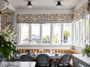 Elegant kitchen with a breakfast nook in front of the kitchen island. This is built around the corner of the room, underneath windows with floral Roman blinds. A dining table is surrounded on two sides by built-in banquette seating, with dining chairs on the other two sides. Marble countertops are visible in the foreground and the dining table is set with cakes and fruit.