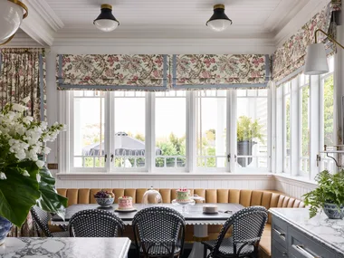 Elegant kitchen with a breakfast nook in front of the kitchen island. This is built around the corner of the room, underneath windows with floral Roman blinds. A dining table is surrounded on two sides by built-in banquette seating, with dining chairs on the other two sides. Marble countertops are visible in the foreground and the dining table is set with cakes and fruit.