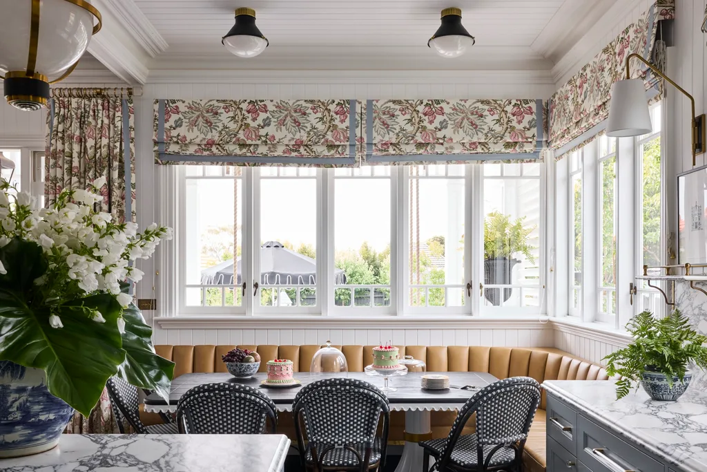 Elegant kitchen with a breakfast nook in front of the kitchen island. This is built around the corner of the room, underneath windows with floral Roman blinds. A dining table is surrounded on two sides by built-in banquette seating, with dining chairs on the other two sides. Marble countertops are visible in the foreground and the dining table is set with cakes and fruit.