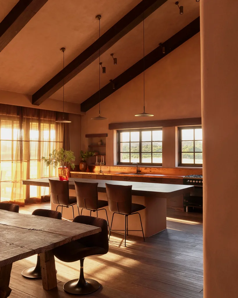 Spacious kitchen with a rustic wooden dining table, modern island, pendant lights, and large windows letting in natural light. Three leather bar stools are positioned in front of the island while three dining chairs are tucked under the dining table. A vaulted ceiling features exposed wooden beams which enhance the rustic vibe.