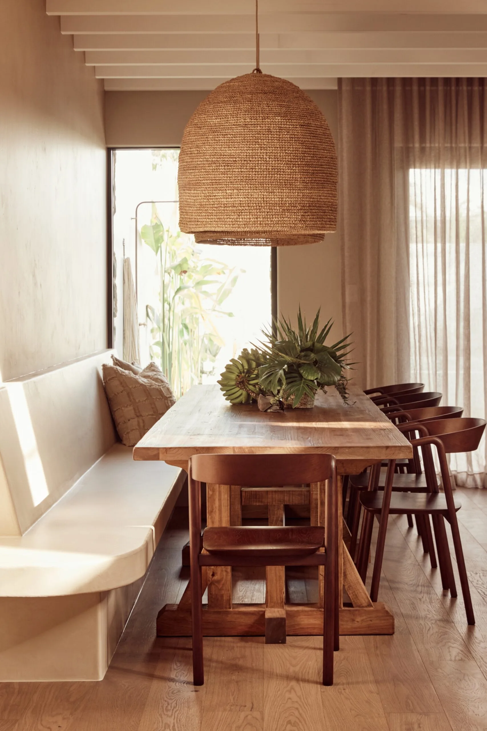 Dining room with wooden table, wooden dining chairs, a a built-in banquette seating, large rattan pendant light, and plants on the table. Natural light through curtains.