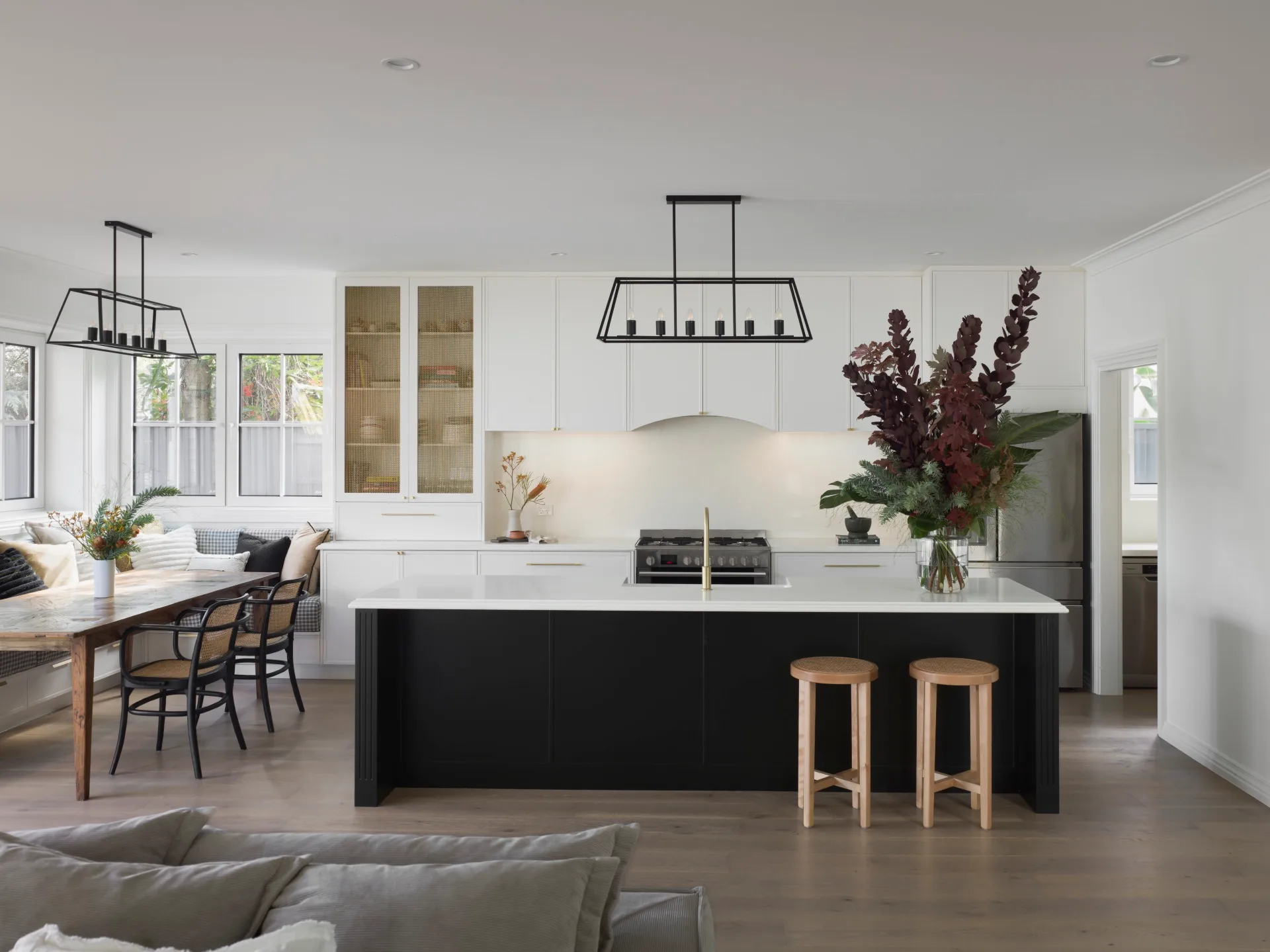 Modern kitchen and dining area with white cabinets, black kitchen island with white benchtop, black metal pendant lights and three vases with flowers adding a pop of colour. To the left, a wooden dining table is surrounded on two sides by built in banquette seating, upholstered in a checkered fabric and decorated with many cushions. On the opposite side of the table two dining chairs are positioned. Two timber bar stools are positioned at the end of the kitchen island, to the right.
