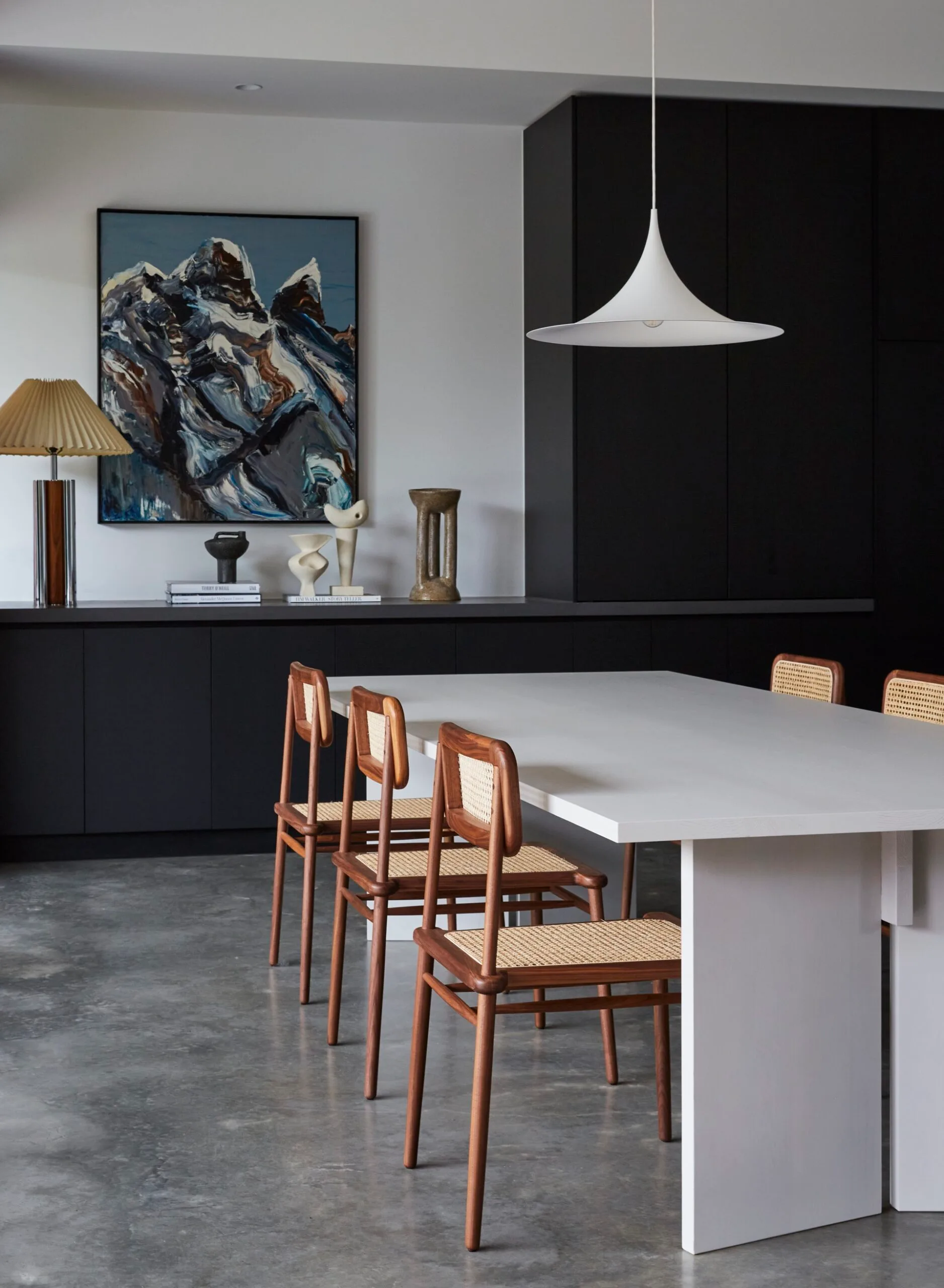 Modern dining room with a white dining table, wooden dining chairs, pendant light, and mountain painting above black cabinetry. The floors are polished concrete.