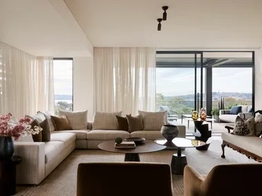 Luxurious living room with beige sectional, pink flowers, round tables, and large windows showcasing a scenic landscape.