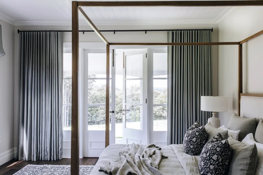 Cozy bedroom with a four-poster bed, decorative pillows, bedside lamp, and large windows with gray curtains.